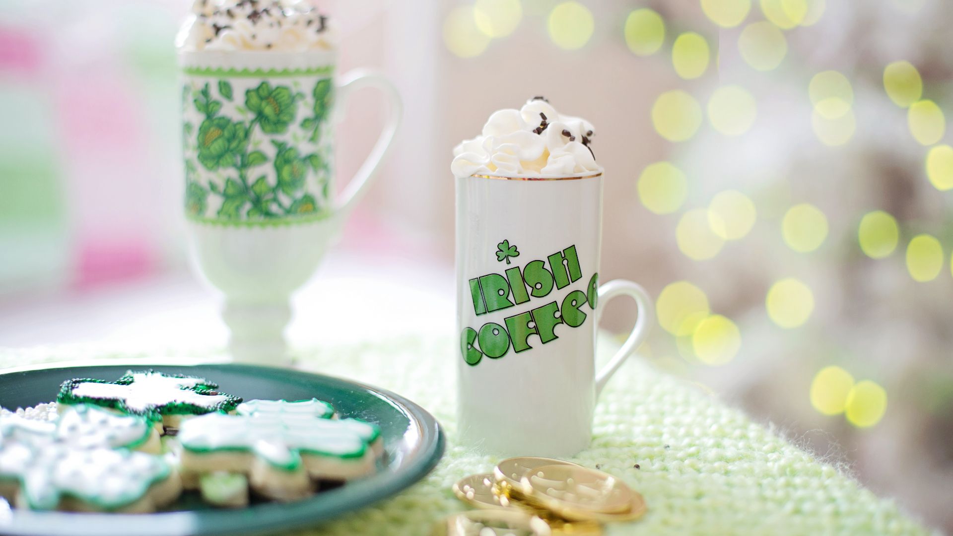 a bokeh background and a table with two cups of coffee and a plate with cookies next to gold coins